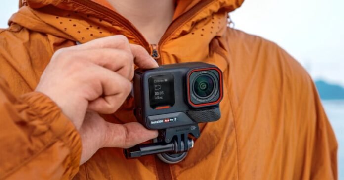 A person in a brown jacket holds an action camera close to his chest. The camera screen displays shooting information. The background is blurred, with the focus on the camera and the hand.