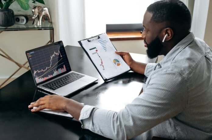 A person holding a clipboard while looking at stock charts on a laptop.