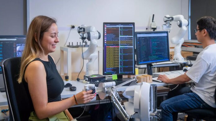 Two people in a laboratory with equipment set up, one touching a mechanical machine and the other working on a computer in the background.